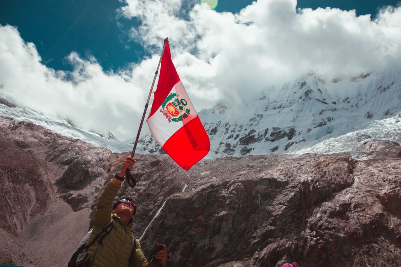 Hiker proudly waves the Peruvian flag against a stunning mountainous background with snow-capped peaks.