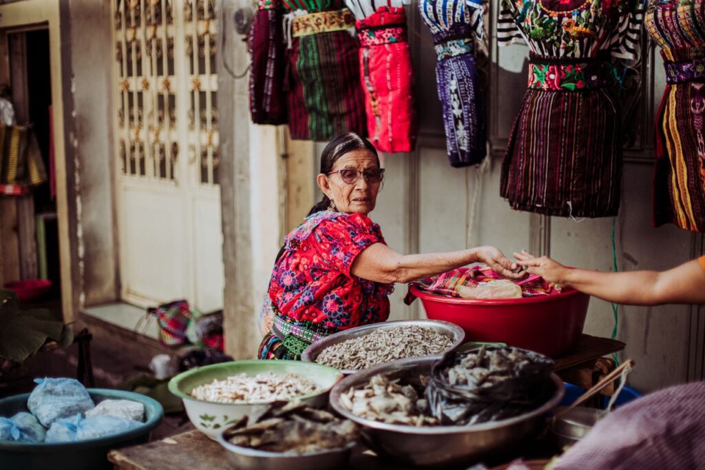 pexels-photo-26088704-2 A female vendor in a vibrant traditional market displaying colorful textiles and local food items.