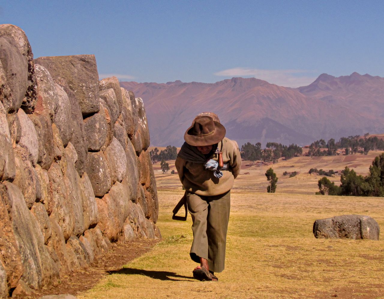 A man in traditional attire walking along a stone wall with mountains in the background.